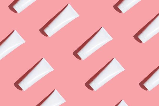 White cream tubes on light pink table. Care about face, hands, legs and body skin. Women beauty products. Cosmetic pattern. Empty place for logo on bottles. Hard light directly above flat lay.