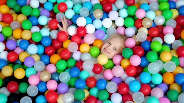 Cute blonde little girl lying on multi coloured plastic balls in big dry paddling pool in playing centre. Smiling at camera. Portrait close up. Having fun in playroom. Leisure Activity.