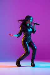 Caucasian female singer portrait isolated on purple studio background in neon light. Beautiful female model in black wear with microphone. Concept of human emotions, facial expression, ad, music, art.