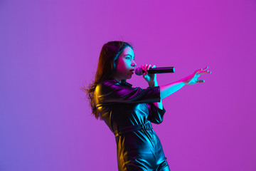 Caucasian female singer portrait isolated on purple studio background in neon light. Beautiful female model in black wear with microphone. Concept of human emotions, facial expression, ad, music, art.