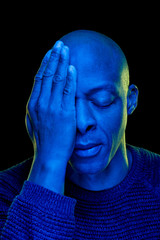 Black man with one eye covered with the hand and the other eye closed. Studio photo with blue light isolated on black background. Desperation concept. Vertical