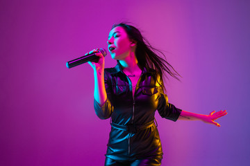 Caucasian female singer portrait isolated on purple studio background in neon light. Beautiful female model in black wear with microphone. Concept of human emotions, facial expression, ad, music, art. - obrazy, fototapety, plakaty