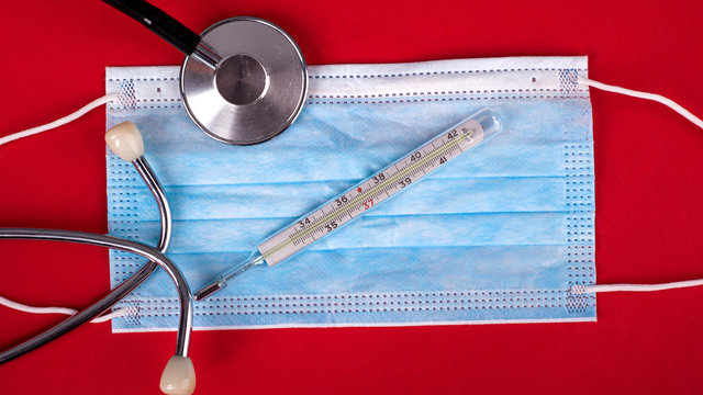 statoscope thermometer and medical disposable mask on a red background top view close-up. Covid-19 influenza, SARS, measles, measles