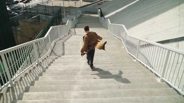Happy Young Man Actively Dancing While Walking down the stairs. He's Wearing a brown long coat. Scene Shot in an Urban Park Next to Business Center. Slow motion. Shooting from the air 
