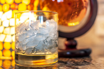 An alcohol glass of crushed ice sits aside a round container of scotch whiskey on a granite countertop with warm light