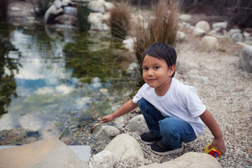 Latino toddler wearing a white tshirt and blue jeans trying to catch insects from a pond using a wodden stick.
