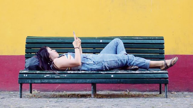 Hispanic woman lay down on the bench texting with a smartphone. 4k