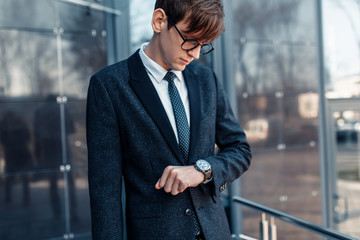Fototapeta premium A young businessman who is late for work looks at the clock on his hand, against the background of a glass office