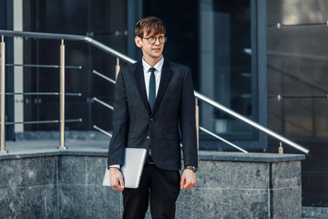 A handsome young businessman holds his laptop while standing in front of a glass building