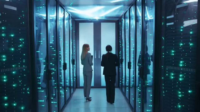 Two women IT server engineers walking through cyberspace corridor, inspecting supercomputer maintenance, coworking in network data center storage.