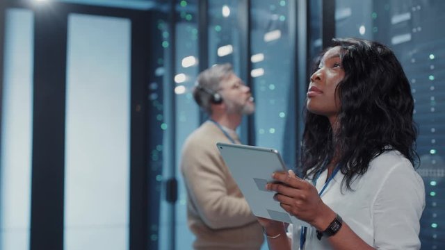Beautiful afro-american IT woman administrator provides deep examination of database storage coworking with technician in futuristic cyber server room. Teamwork, business concept.