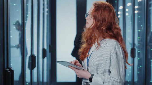 Red-haired attractive IT woman visually examines row of server racks typing data cloud information into tablet collaborating with man in secure data center.