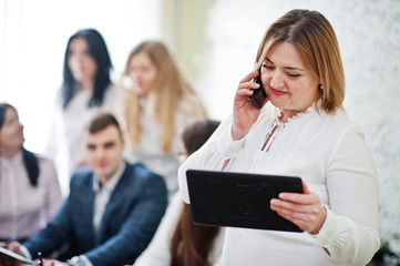 Obraz premium Portrait of caucasian woman in white blouse hold tablet and speak on phone against business people group of bank workers.
