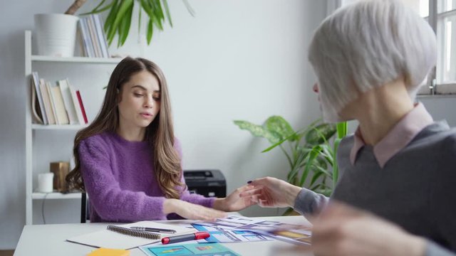 Female designer sitting at table with open laptop, turning to busy colleague, clarifying details of project and discussing webiste interface templates printed on papers in office