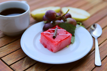Food background. Delicious breakfast. In the center of the frame on a wooden table is a white plate with a slice of sponge cake with red jelly, fruits and a cup of coffee. Close-up side view.