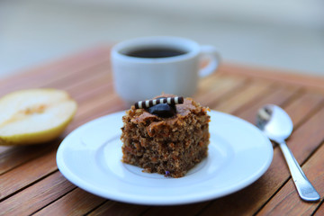 Blurred food background. On a wooden table is a white plate with a slice of chocolate cake and a spoon, in the background a cup of coffee and fruit. Close-up, side view, horizontal. Food concept.