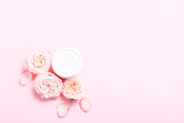 Open jar with cream for skin care and roses flowers around on pink background.