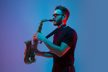 Young caucasian jazz musician playing the saxophone on gradient blue-purple studio background in neon light. Concept of music, hobby, festival. Joyful attractive guy. Colorful portrait of artist. - obrazy, fototapety, plakaty