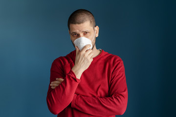 POrtrait of adult caucasian man standing in front of the blue wall wearing red sweater and protective mask on his face against virus flue bacteria protection health issues front view