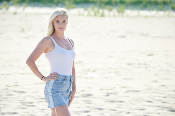 Stunning beautiful woman with blonde hair poses on beach in white tank top and denim skirt