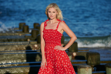 Stunning young blonde haired woman poses in red sundress on the beach - wooden pier pilings behind model