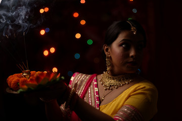 Close up portrait of young and beautiful Indian Bengali woman in Indian traditional dress celebrating Diwali with incense sticks and flowers in dark background.Indian lifestyle and Diwali celebration.