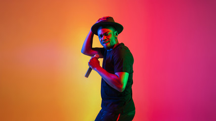 Young african-american musician singing on gradient orange-purple studio background in neon light. Concept of music, hobby, festival. Joyful party host, stand upper. Colorful portrait of artist.