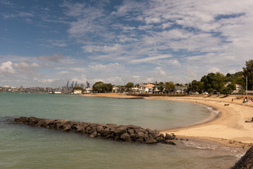 The sandy beach at Torpedo Bay in Devonport, with the Auckland City skyline visible across the Waitemata Harbour.