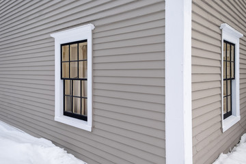 Two horizontal opening vintage windows in a tan colored wooden building. The windows have white trim around them. The exterior wall of the building is a beige clapboard. A snow drift is on the ground.