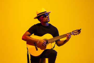 Young and joyful african-american musician playing guitar and singing on gradient orange-yellow...