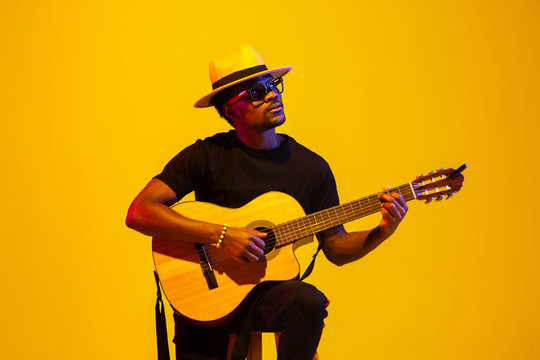 Young and joyful african-american musician playing guitar and singing on gradient orange-yellow studio background in neon light. Concept of music, hobby, festival. Colorful portrait of modern artist.