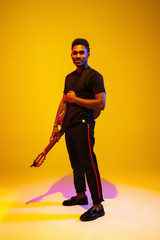 Young and joyful african-american musician playing guitar and singing on gradient orange-yellow studio background in neon light. Concept of music, hobby, festival. Colorful portrait of modern artist.