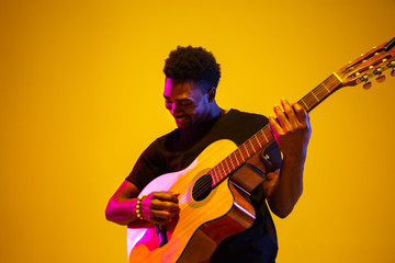 Young and joyful african-american musician playing guitar and singing on gradient orange-yellow studio background in neon light. Concept of music, hobby, festival. Colorful portrait of modern artist.