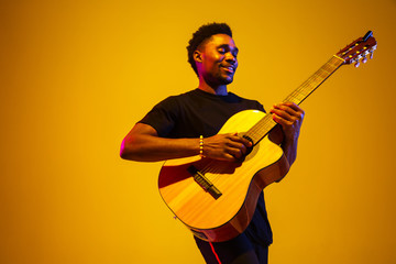 Young and joyful african-american musician playing guitar and singing on gradient orange-yellow studio background in neon light. Concept of music, hobby, festival. Colorful portrait of modern artist. - obrazy, fototapety, plakaty