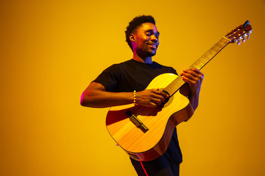 Young and joyful african-american musician playing guitar and singing on gradient orange-yellow studio background in neon light. Concept of music, hobby, festival. Colorful portrait of modern artist.