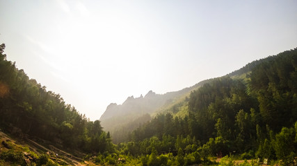 Green valley in the mountains. Trees and grass on the slopes. Rock ledge, people and wooden buildings in the distance. Bright sunlight, glare. Summer Sunrise Concept.