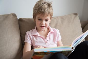 the child is reading a large book, sitting comfortably on the sofa.