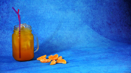 A glass mug of citrus juice with ice and a straw with pieces of mandarin next to it stands on a blue background. © Алексей Желтухин