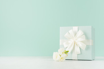 Obraz na płótnie Canvas Easter festive background in trendy green mint menthe color on white wood table - fresh white flowers, elegant standing square gift box with knot.
