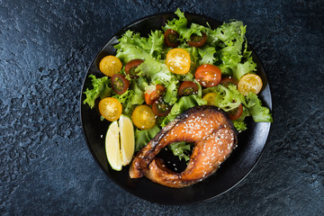 Salmon steak fried in teriyaki sauce with fresh lettuce and tomatoes on a black background. Top view, close-up