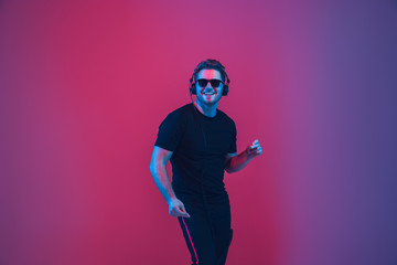 Obraz na płótnie Canvas Young caucasian musician in headphones singing on gradient pink-purple background in neon light. Concept of music, hobby, festival. Joyful party host, DJ, stand upper. Colorful portrait of artist.
