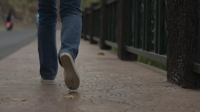 View from rear side of asian woman legs wearing jeans walking leisurely on the footpath along the road in the evening. Close up follow shot with low angle view. Slow motion.