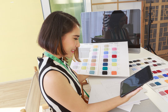 Smiling beautiful Asian woman fashion designer with tape measure holding color sample and looking tablet in workplace at home - Small business owner and SME concept
