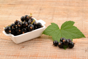 Black currant in white bowl, on wooden background. Fruits and green leaves.