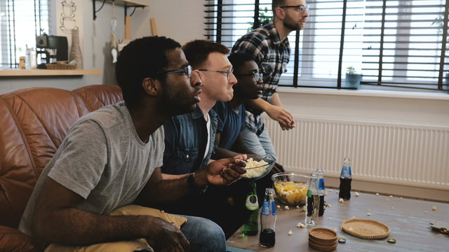 Mixed race male supporters watch sports on TV 4K. Fans celebrate goal victory holding British flag cushion. Slow motion. - Powered by Adobe