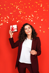 Portrait of young beautiful woman in marsala pantsuit with present box on red background with sparkles.