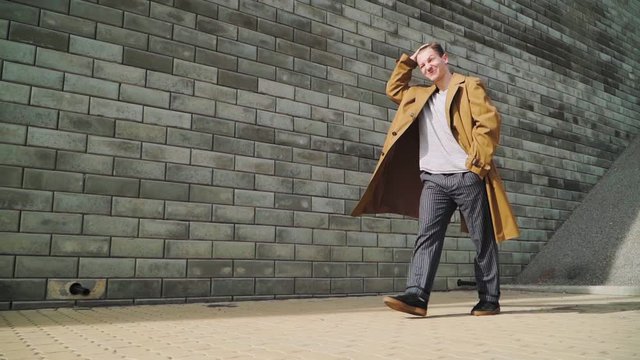 Stylish Beautiful Young Happy Caucasian Guy Goes along the City Wall and smiles. He is dressed in a Brown Long Coat. Slow motion. Close up. shooting on Steadicam - Powered by Adobe