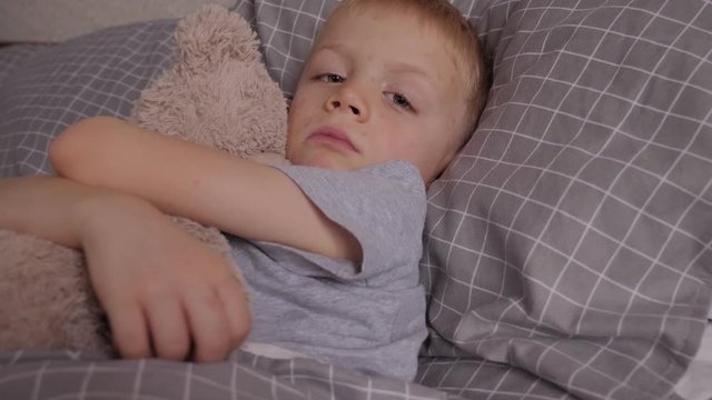 A little five-year-old boy with chicken pox is lying in bed with his favorite Teddy bear. A child with inflamed skin and blisters from chickenpox is lying in bed.