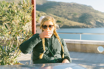 Beautiful young woman enjoys coffee at a sea side cafe with blonde braids sipping mug
