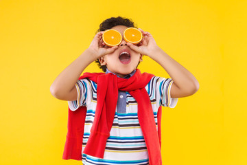 Emotional dark skinned curly boy in blue striped T-shirt, red hoodie is screaming on bright yellow background. Fashionable black child is holding oranges like glasses. Modern children concept.